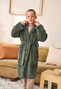 Green, plush, hooded robe with a belt, featuring two front pockets and a soft texture, worn by a person in a cozy indoor setting.