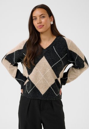 Femme aux longs cheveux foncés porte un pull en laine noir et beige à motif losanges et un pantalon noir, debout les mains sur les hanches devant un fond uni.