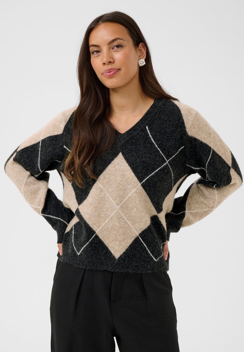 Femme aux longs cheveux foncés porte un pull en laine noir et beige à motif losanges et un pantalon noir, debout les mains sur les hanches devant un fond uni.