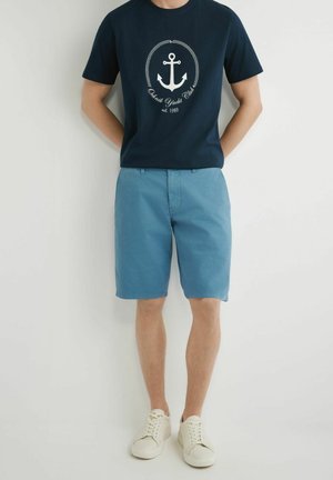 Mężczyzna w granatowej koszulce z logo kotwicy, jasnoniebieskich szortach i białych trampkach, stojący na jednolitym jasnym tle.