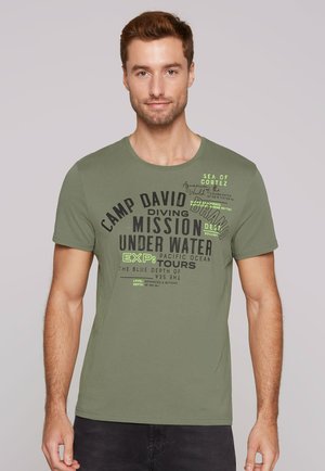 Young man wearing olive green T-shirt with black and neon green scuba diving text and graphics, standing against plain light background.