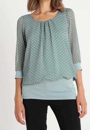 Femme portant un chemisier bleu clair à pois avec des manches trois-quarts transparentes et une sous-couche bleu clair unie, associée à un pantalon noir.