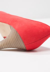 Chaussure à talon haut en daim rouge avec un bout pointu et un talon en bois effilé présentant une surface texturée.
