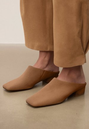 Jalad kannavad beežikaspruune mugavaid madala plokkingaga poolsaapad, mis on kombineeritud sobivate beežide laiade pükstega, kõnniteekorralisel siledal põrandal.