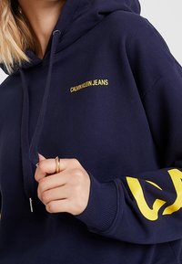 Marinblå luvtröja med gul "Calvin Klein Jeans"-logotyp, utrustad med dragsko i huvan och ribbade muddar. Mjuk tygstruktur.
