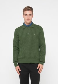 Pull en maille torsadée vert avec un col, comprenant des poignets et un ourlet côtelés, et un petit logo sur la poitrine. Porté par-dessus une chemise bleue.