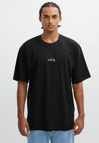 Joven con cabello trenzado que lleva una camiseta negra holgada con el texto blanco "Le Petit Café" y jeans de color azul claro, de pie contra un fondo blanco.