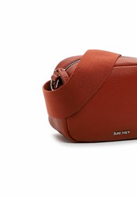 Väike põlenud oranži nahast õlakott laia kangastriibu ja hõbedase luku ning esiküljel metallist kaubamärgi logoga "SURI FREY".