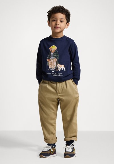 Marineblaues Langarmshirt mit einem Bärengrafik, khakifarbene Hose und marineblaue Sneaker mit weißen Akzenten. Kind steht mit den Händen in den Taschen.