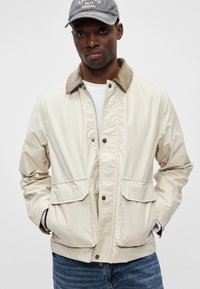 Ανοιχτό μπεζ σακάκι με κολάρο από καπιτονέ, δύο μπροστινές τσέπες και κλείσιμο με φερμουάρ/κουμπί, συνδυασμένο με λευκό πουκάμισο και μπλε τζιν.