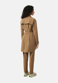 Tan trenchcoat med plisseringer og bælte, parret med matchende bukser. Frakken har en flapdetalje bagpå. Letvægtsstof, casual sneakers.