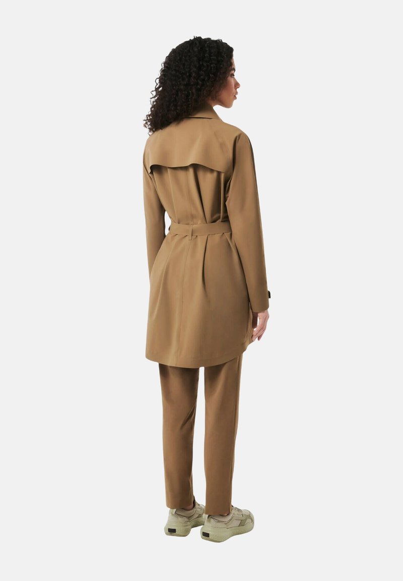 Tan trenchcoat med plisseringer og bælte, parret med matchende bukser. Frakken har en flapdetalje bagpå. Letvægtsstof, casual sneakers.