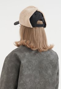 Black denim cap with beige fluffy lining, featuring a rear adjustable strap. The model wears a textured gray jacket with detailed embroidery.