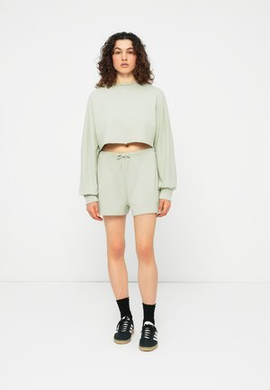 Femme aux cheveux bouclés portant un sweat court vert clair et un short assorti, associés à des chaussettes noires et des baskets noires à rayures blanches.