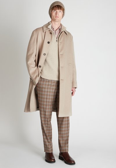 Beige wollen overcoat met grote knopen, gebreide crèmekleurige trui en bruine geruite broek. Afgemaakt met bruine leren laarzen en een gebreide beanie.