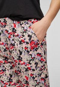 Main dans la poche d'un pantalon à taille élastique avec un motif floral rouge, noir et violet, associé à un haut noir.