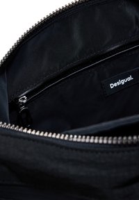Interno di una borsa nera con zip, dalla texture liscia, caratterizzato da un design compartimentato e un'etichetta di marca ben visibile, "Desigual."
