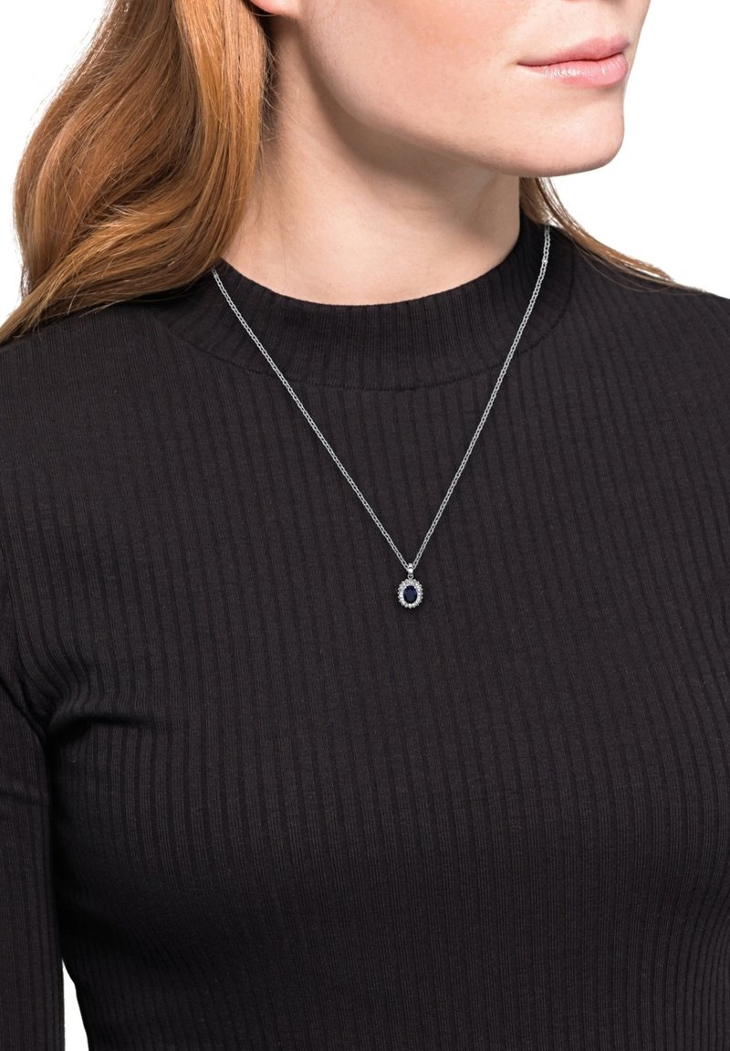 Srebrny naszyjnik z łańcuszka z owalnym niebieskim kamieniem otoczonym okrągłym skupiskiem przezroczystych kamieni, noszony na fakturowanej czarnej bluzce.