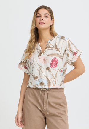 Kobieta o długich blond włosach nosi beżową, kwiecistą bluzkę z krótkim rękawem oraz jasnobrązowe spodnie ze sznurkiem w talii na jednolitym tle.