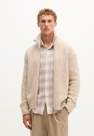 Junger Mann trägt einen beigen grob gestrickten Reißverschluss-Pullover über einem hell karierten Hemd und beigen Hosen, steht vor einfachem Hintergrund.