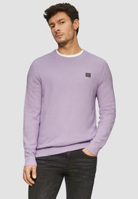 Helles lila strukturiertes Sweatshirt mit Rundhalsausschnitt, gerippten Bündchen und einem kleinen schwarzen Logo-Patch auf der Brust. Kombiniert mit dunklen Jeans.