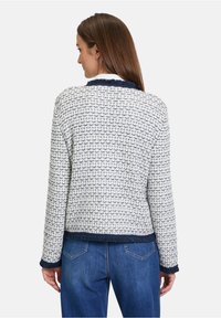 Texturierte weiße und marineblaue Jacke mit geometrischem Muster. Verfügt über einen marineblauen ausgefransten Kragen und Manschetten, kombiniert mit blauen Jeans.