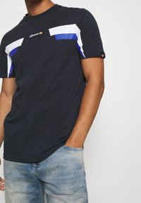 Mężczyzna w czarnej koszulce ellesse z białymi i niebieskimi paskami na klatce piersiowej oraz jasnoniebieskich dżinsach, ręka partially w kieszeni.