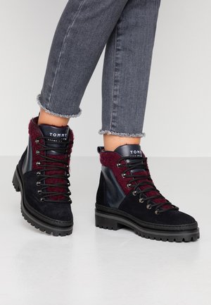 Sorte og bordeauxfarvede snørebånds-ankelstøvler med tykke såler, båret med flossede grå jeans på lys gulv.