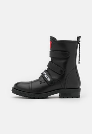 Černá kožená kotníková bota s třemi nastavitelnými pásky, kulatou špičkou, robustní podrážkou a malým červeným srdíčkem poblíž horní části.