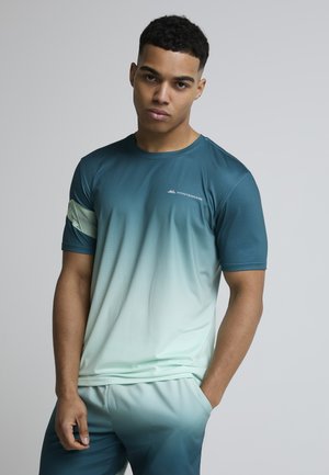 Junger Mann steht vor einfachem Hintergrund, trägt ein Sportshirt und Shorts in Türkis- und Hellgrünverlauf, Hände in den Taschen, schaut in die Kamera.