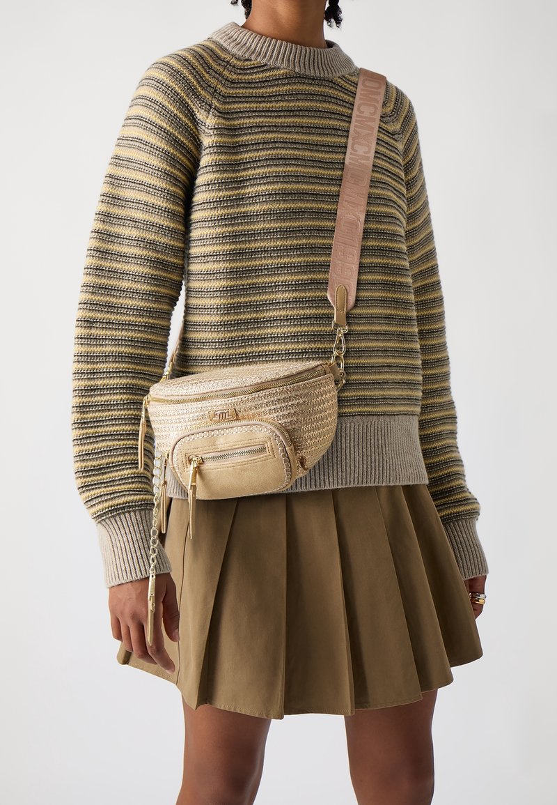 Personne portant un pull rayé et une jupe plissée marron, portant un sac bandoulière tissé beige avec une large sangle de marque.