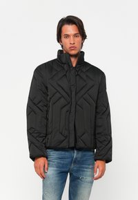 Sort quiltet pufferjakke med høj krave, lynlås og geometrisk tekstur. Båret over blå slidte jeans.