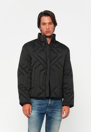 Black quilted puffer jacket with high collar, zip closure, and geometric texture. Worn over blue distressed jeans.