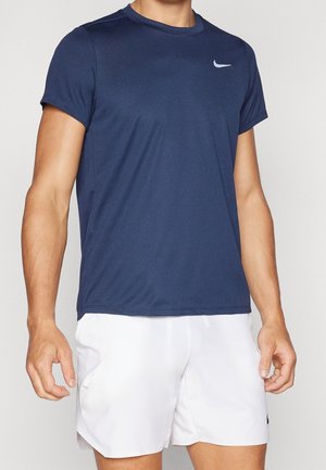 Mand iført navyblå Nike t-shirt med korte ærmer og hvide sportsshorts, står med armene afslappet ned langs siderne.