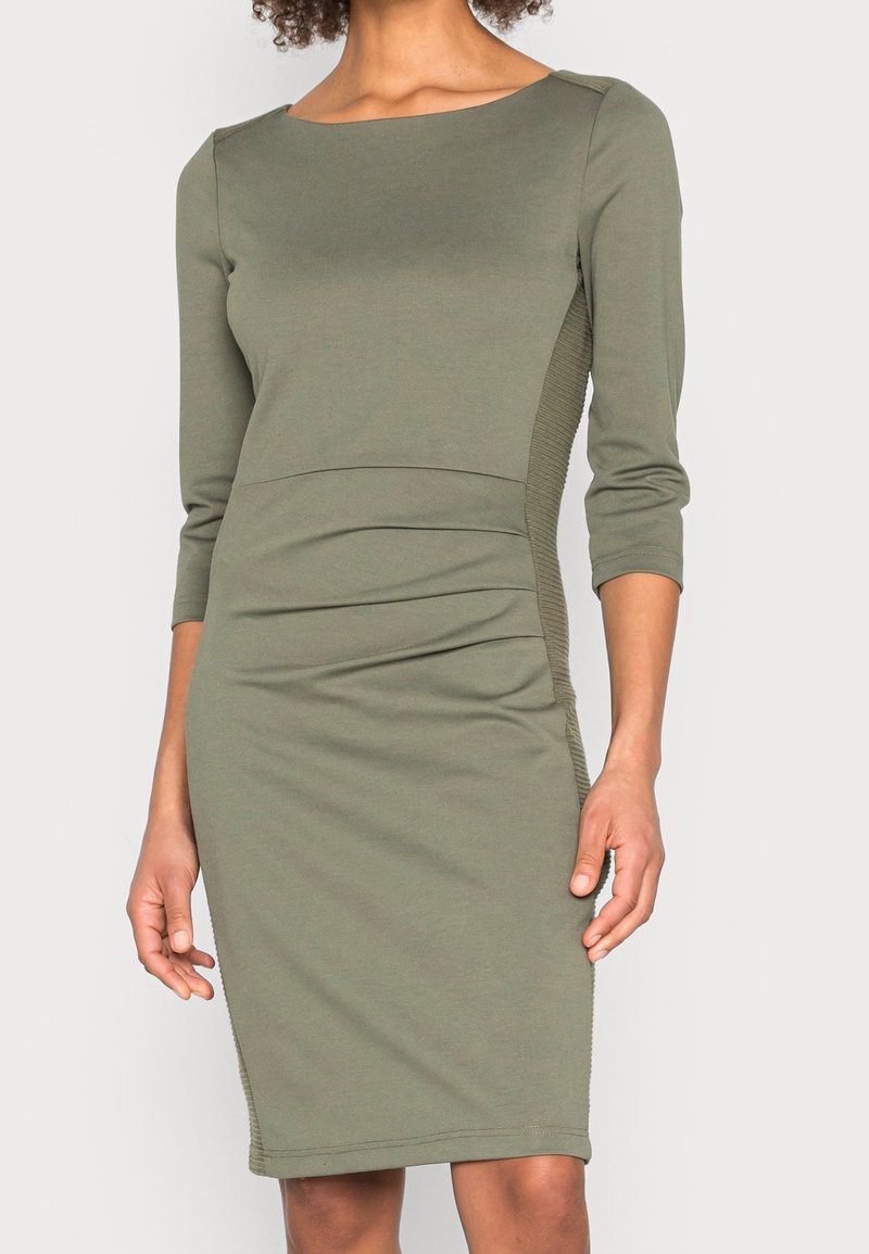 Femme portant une robe olive longueur genou avec manches trois-quarts et panneaux latéraux côtelés, debout devant un fond clair uni.