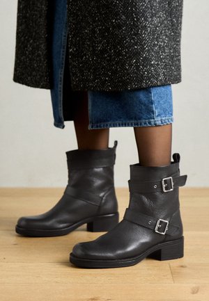 Bottines en cuir noir à doubles boucles portées avec des collants noirs transparents, une jupe en jean bleue et un manteau foncé texturé.