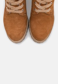 Hnědé semišové kotníkové boty s kulatou špičkou, texturovaným povrchem a kontrastními šňůrkami. Šití podél podrážky dodává strukturu.