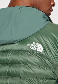 Doudoune verte à finition mate, dotée d'une capuche, de coutures horizontales et du logo blanc The North Face sur le côté gauche.