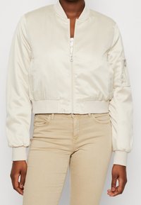 Kobieta w kremowej satynowej kurtce bomber z zamkiem błyskawicznym z przodu oraz beżowych dopasowanych spodniach, stojąca na jednolitym tle.