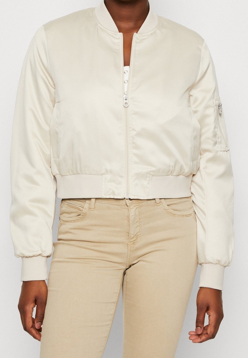 Kobieta w kremowej satynowej kurtce bomber z zamkiem błyskawicznym z przodu oraz beżowych dopasowanych spodniach, stojąca na jednolitym tle.