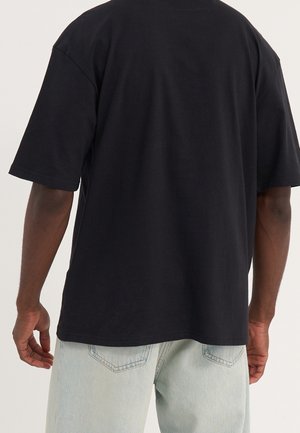 Mann trägt ein schlichtes schwarzes T-Shirt mit kurzen Ärmeln und hellblauen Jeans, von hinten vor einem einfarbigen Hintergrund gezeigt.