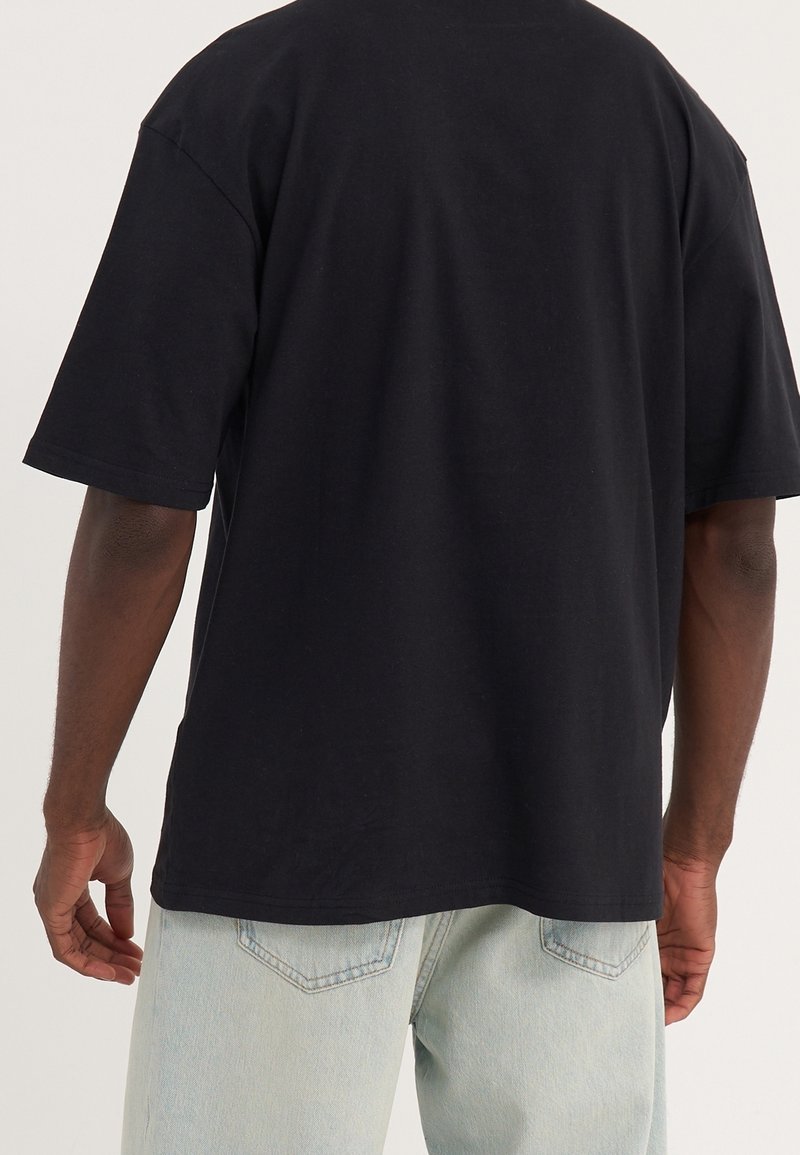 Homme portant un t-shirt noir uni à manches courtes et un jean bleu clair, vu de dos sur un fond uni.