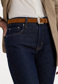 Diepblauwe denim jeans met een slim fit, voorzien van koperen stiksel, voorzakken en een bruine suède riem met een zilveren gesp.