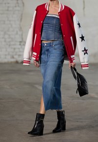 Modelo vistiendo una chaqueta varsity roja y blanca sobre un top de tubo de denim, una larga falda de denim con abertura, botas negras hasta el tobillo y sosteniendo un bolso negro.