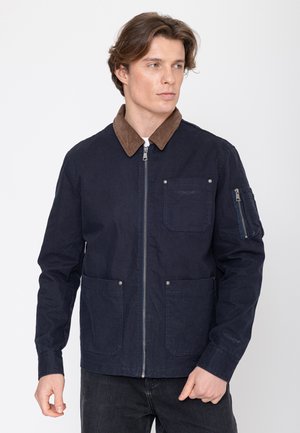 Jonge man zijwaarts kijkend, gekleed in een marineblauw jack met een bruine corduroy kraag, voorzakken en een ritssluiting op de linkermouw.