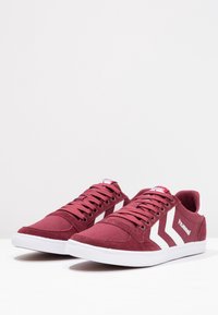 Para bordowych sneakersów Hummel z białymi chevronowymi paskami i białymi podeszwami, umieszczona na jednolitym białym tle.