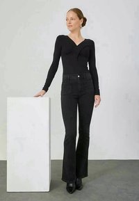 Mulher com uma blusa preta de manga longa e jeans flare pretos de cintura alta encosta-se a um pedestal retangular branco, olhando para o lado.
