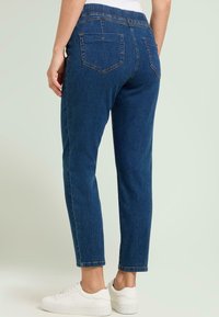 Vaqueros de mezclilla azul con pierna cónica, cintura elástica y bolsillos traseros, combinados con zapatillas blancas. La tela tiene una textura suave.