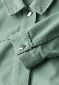 Lichtgroene denim jas met een kraaghals, zilveren drukknoop, gestructureerde stof en gestikte details op de schouders en mouwen.