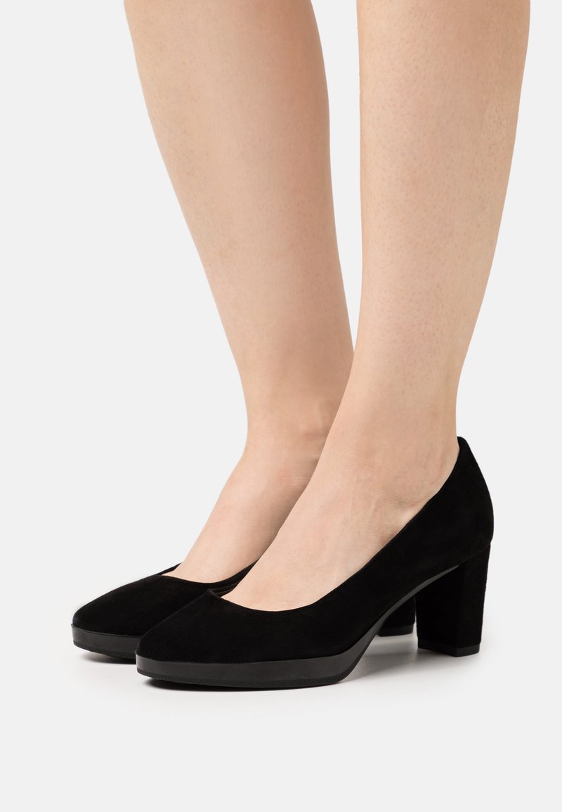 Jambes de femme portant des chaussures basses à talons carrés en daim noir sur fond blanc, montrant les vues latérale et frontale des chaussures.